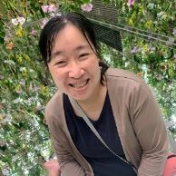 Emiko Tanaka smiling, with shoulder-length dark hair and a beige cardigan, in front of a wall of green plants.