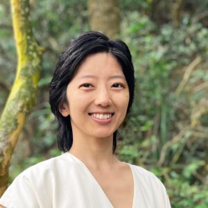 Lily Zhang smiling, with chin-length black hair and a white top, standing among trees and green undergrowth.