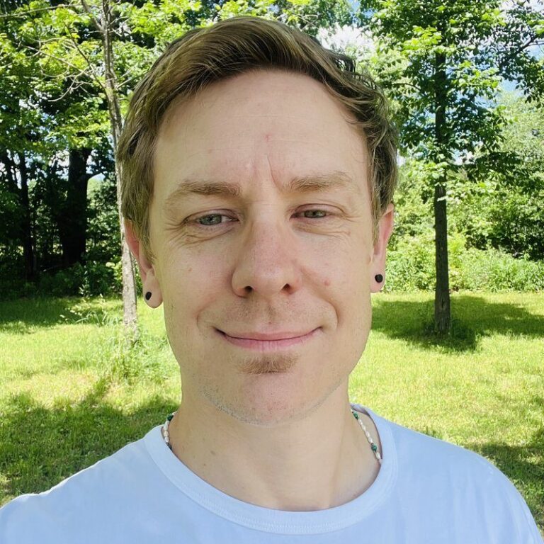 Luke Smith smiling outdoors in a white t-shirt, with green trees and grass behind him.
