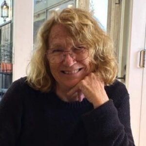 Marina Bagley smiling, with curly fair hair and glasses, resting her chin on her hand at a cafe table