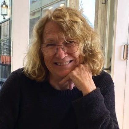 Marina Bagley smiling, with curly fair hair and glasses, resting her chin on her hand at a cafe table