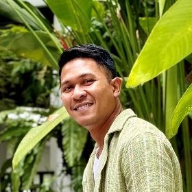 Praphat Manuelo Ruengthanoo, smiling, in a green short-sleeved shirt in front of large tropical leaves.