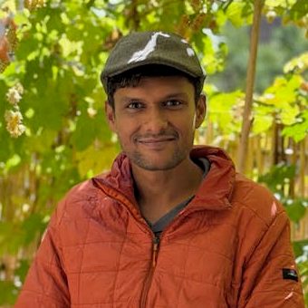 Rahul Tola, smiling, in a grey flat cap and orange quilted jacket in front of climbing hop vines and a bamboo fence.