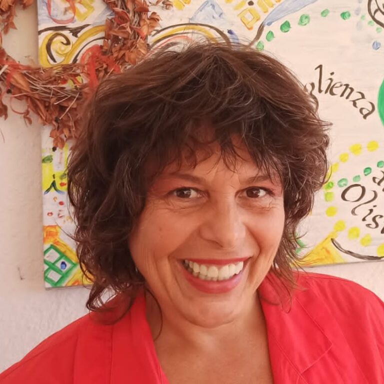 Ramona Bavassano, smiling, with short dark curly hair and a red jacket, in front of a painted permaculture mural.