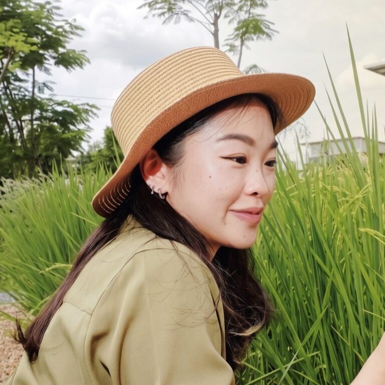 Santhila Chanoknamchai in a straw hat and olive shirt, face turned to the side, beside tall green rice plants in a field.