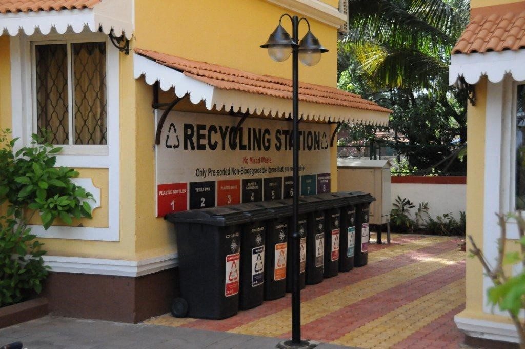 A row of black recycling bins under a Recycling Station sign beside a yellow building in Goa.