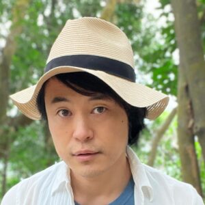 Wataru Shinji in a straw hat with a black band, a white shirt over a blue T-shirt, standing among trees.