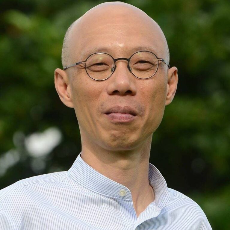 Wong Kam Sing, bald, in round wire-rimmed glasses and a striped collarless shirt, outdoors against green foliage.