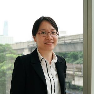 Woraporn Poonyakanok, wearing glasses and a dark blazer over a white shirt, smiling beside a window.